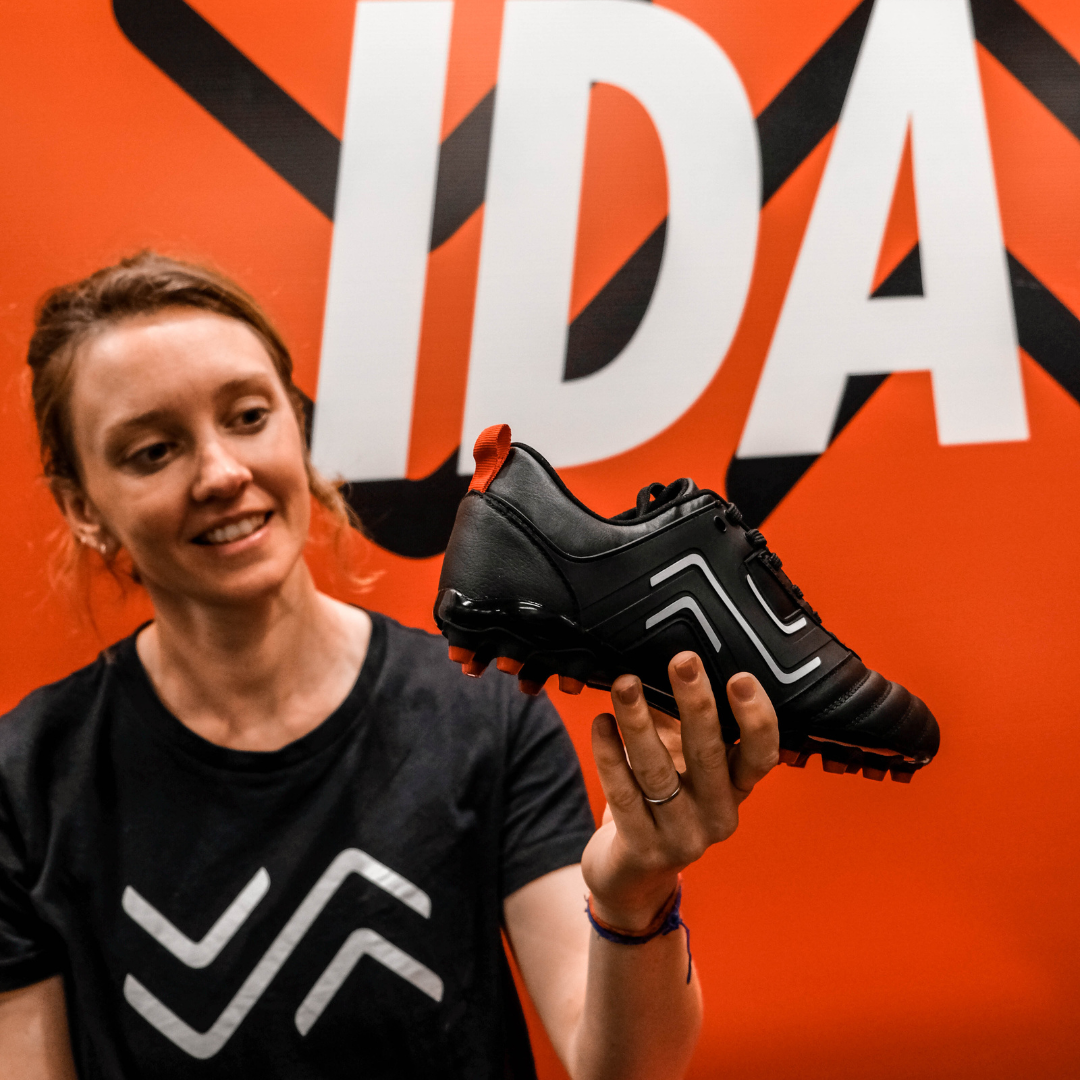 IDA Sports co-founder Laura Youngson sits in front of a red wall holding the IDA Classica black leather women's soccer cleat.