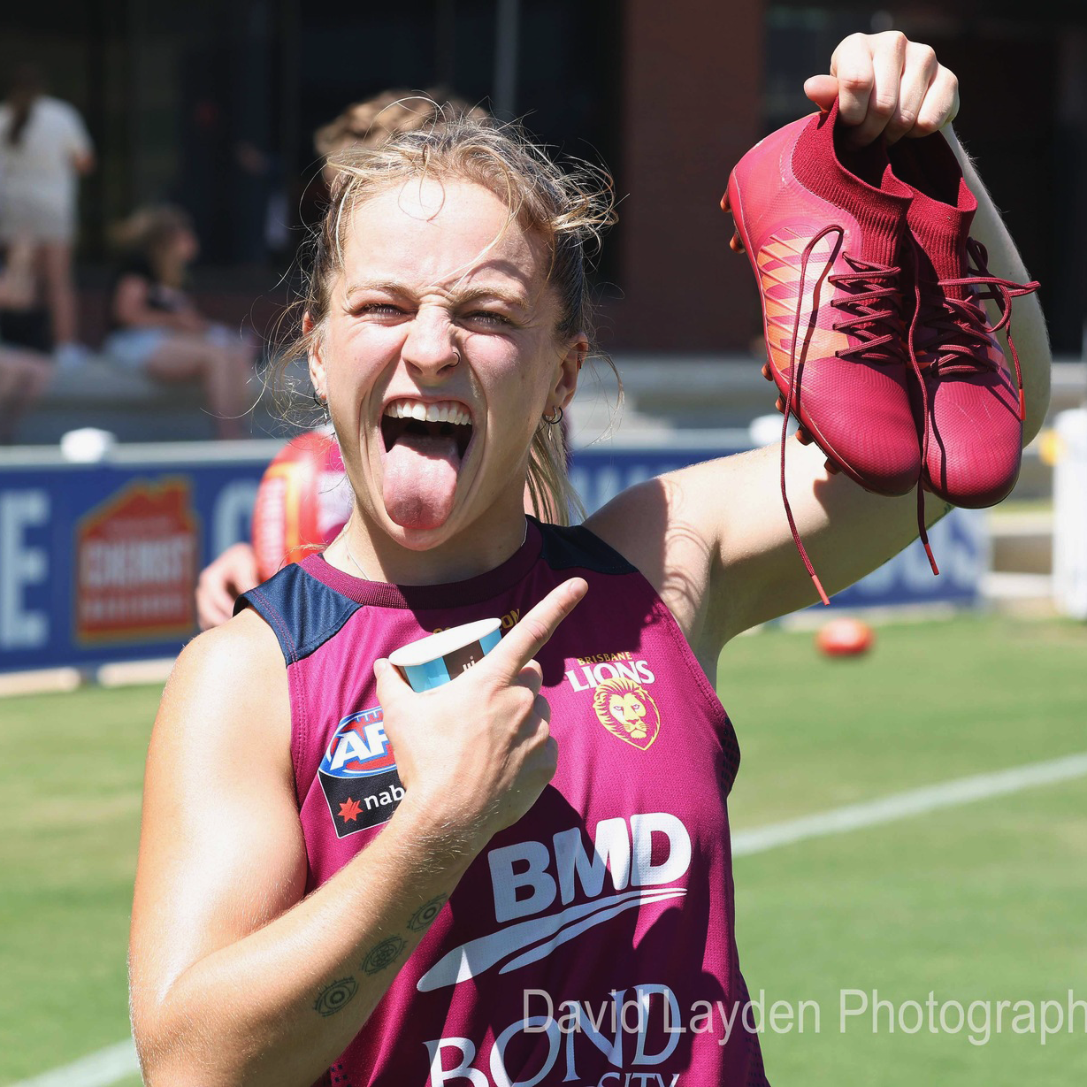 Belle Dawes AFLW player for the Brisbane Lions holds a pair of the burgundy IDA Rise RG/AG boots while smiling.
