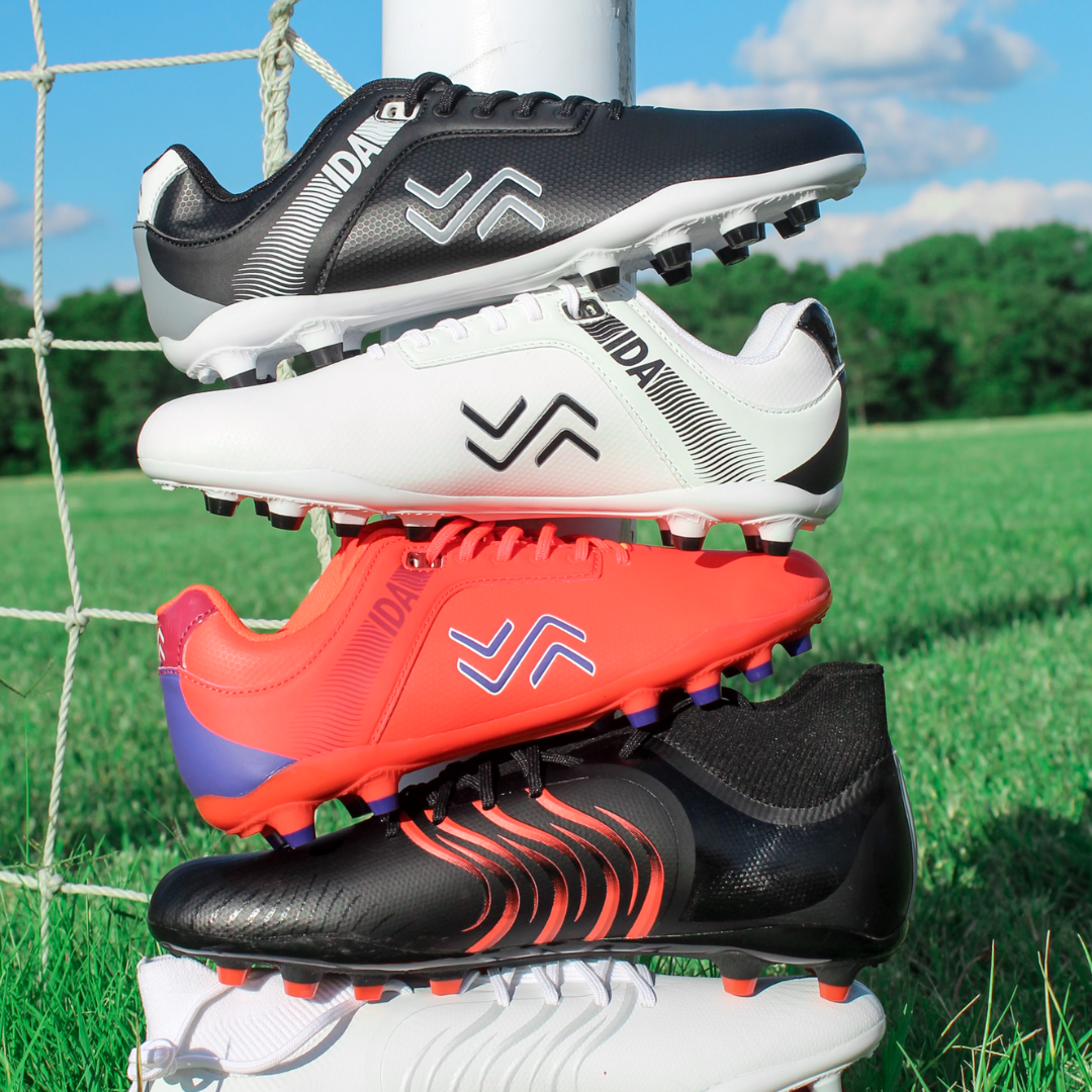 IDA women's soccer cleats are stacked on top of each other on a soccer field next to a soccer goal net.