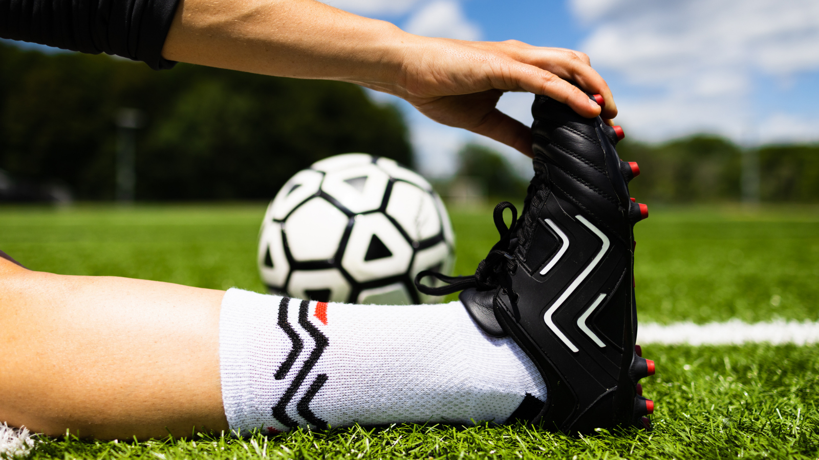 player sits on football pitch with outstretched leg and reaches for toes to stretch while wearing Ida Sports football boots