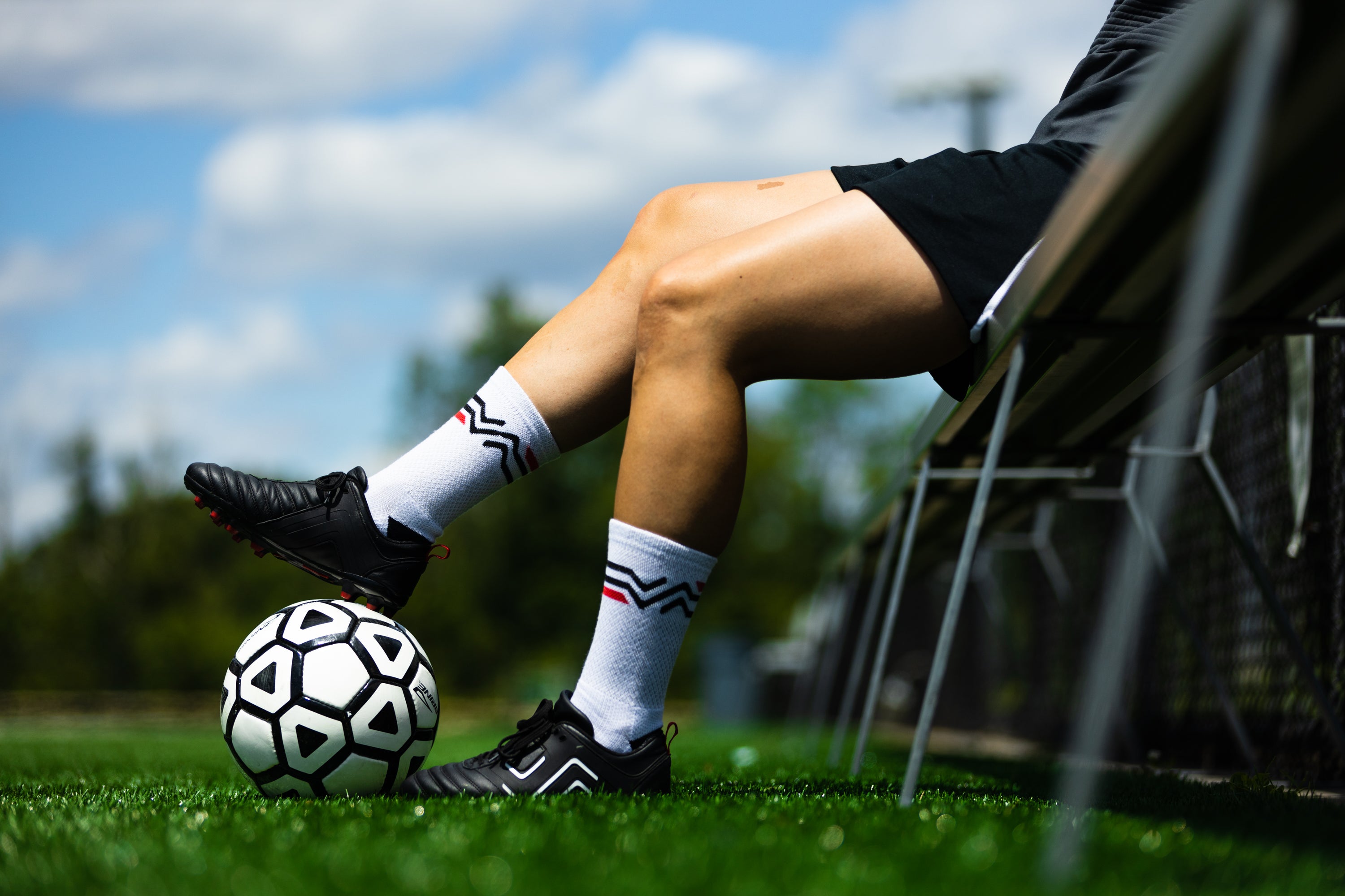 Female football player wearing the IDA Classica black women's football boots sits on a bench next to a football pitch, with right foot balanced on a black and white football.